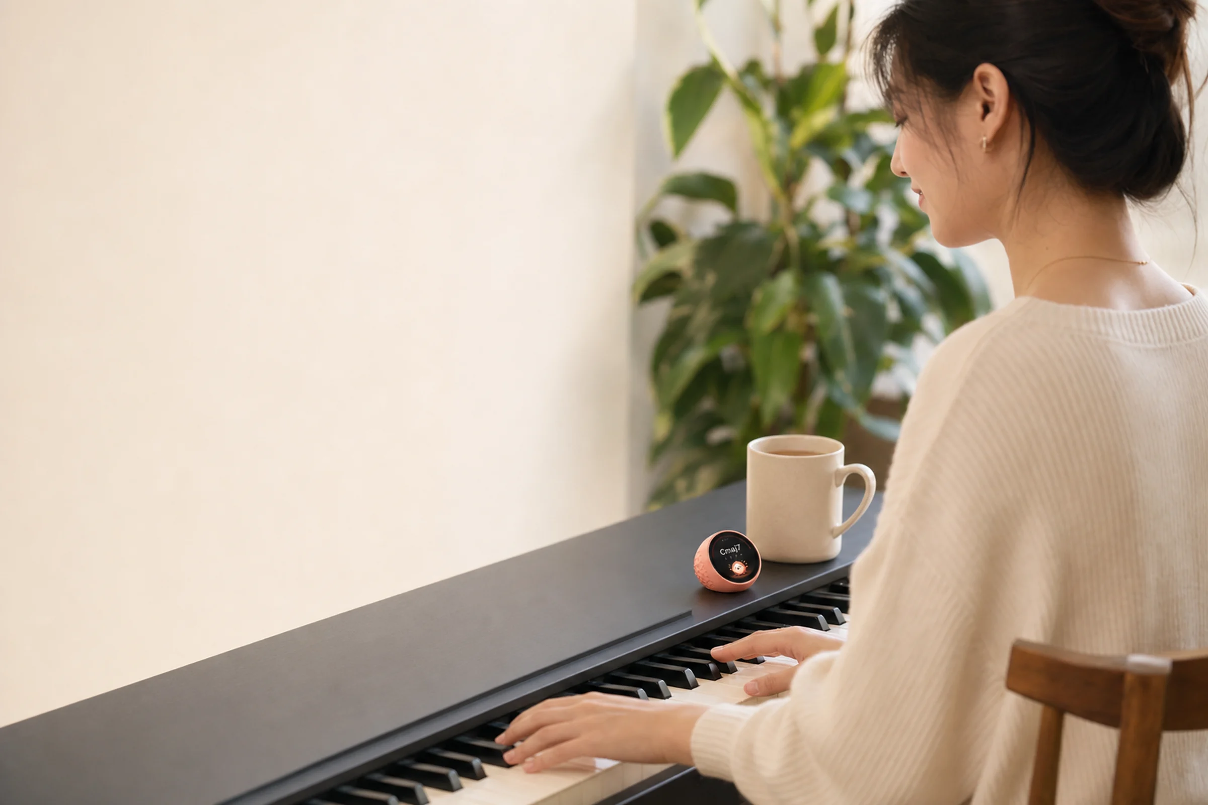A woman playing a digital piano. A tiny coral Keetchi sits on the piano next to her mug, showing Cmaj7.